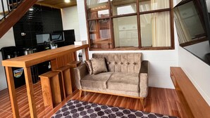 A loft-style living area with a wooden staircase, a sofa, a dining table, and a kitchen in the background. - Sanctuary House (Dieng Kulon)