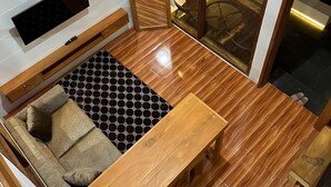 A modern room with a wooden floor, a sofa, a table, a TV mounted on the wall, and a large window with curtains. - Sanctuary House (Dieng Kulon)