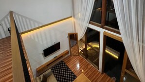 A loft-style bedroom with a wooden ceiling, a built-in bed, a small seating area, and a large window with curtains. - Sanctuary House (Dieng Kulon)