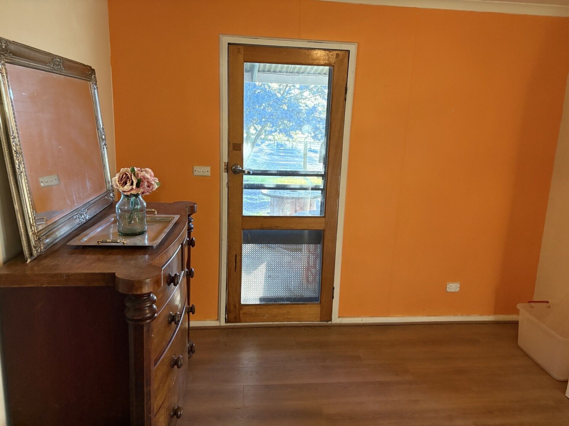 A room with a wooden dresser, a door leading outside, and a vase with flowers on the dresser.