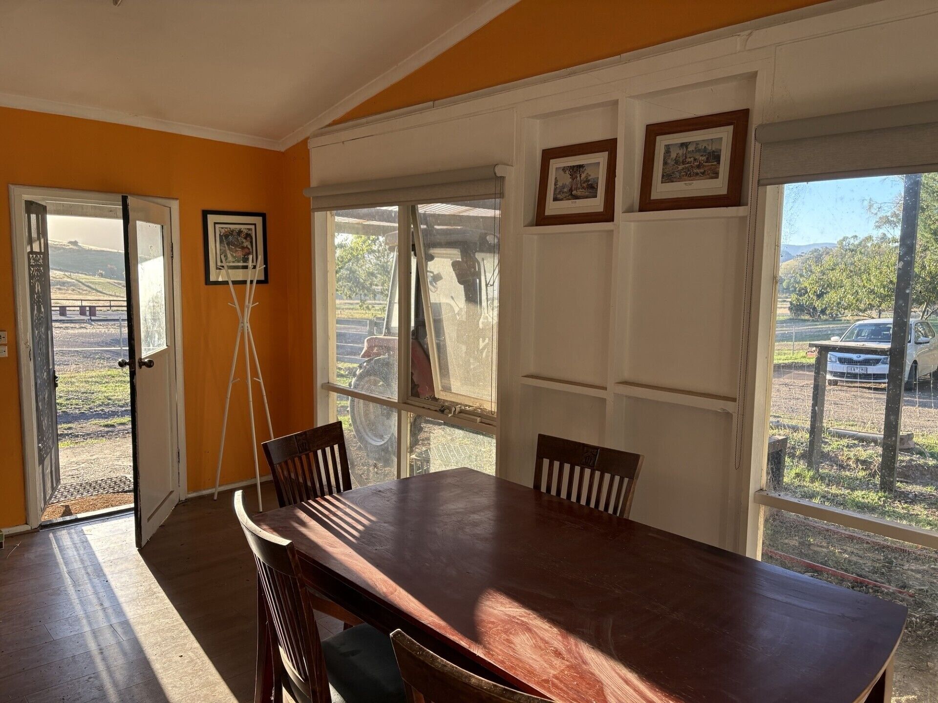 A dining area with a wooden table and chairs, a window with a view of a parking lot, and framed pictures on the wall.