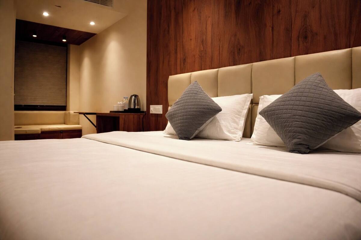 A hotel room with a large bed, a wooden headboard, a bedside table with a lamp, and a window with blinds.