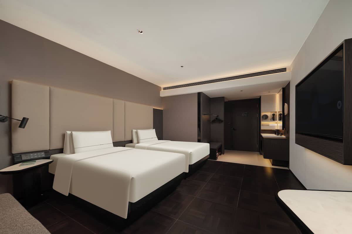 A modern hotel room with two beds, a large mirror, a TV, and a bathroom with a sink and mirror.
