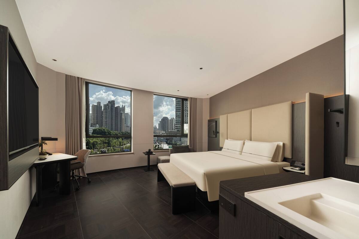 A modern hotel room with a large bed, a desk, a chair, a bench, a small table, a nightstand, a large window with a city view, and a bathtub.