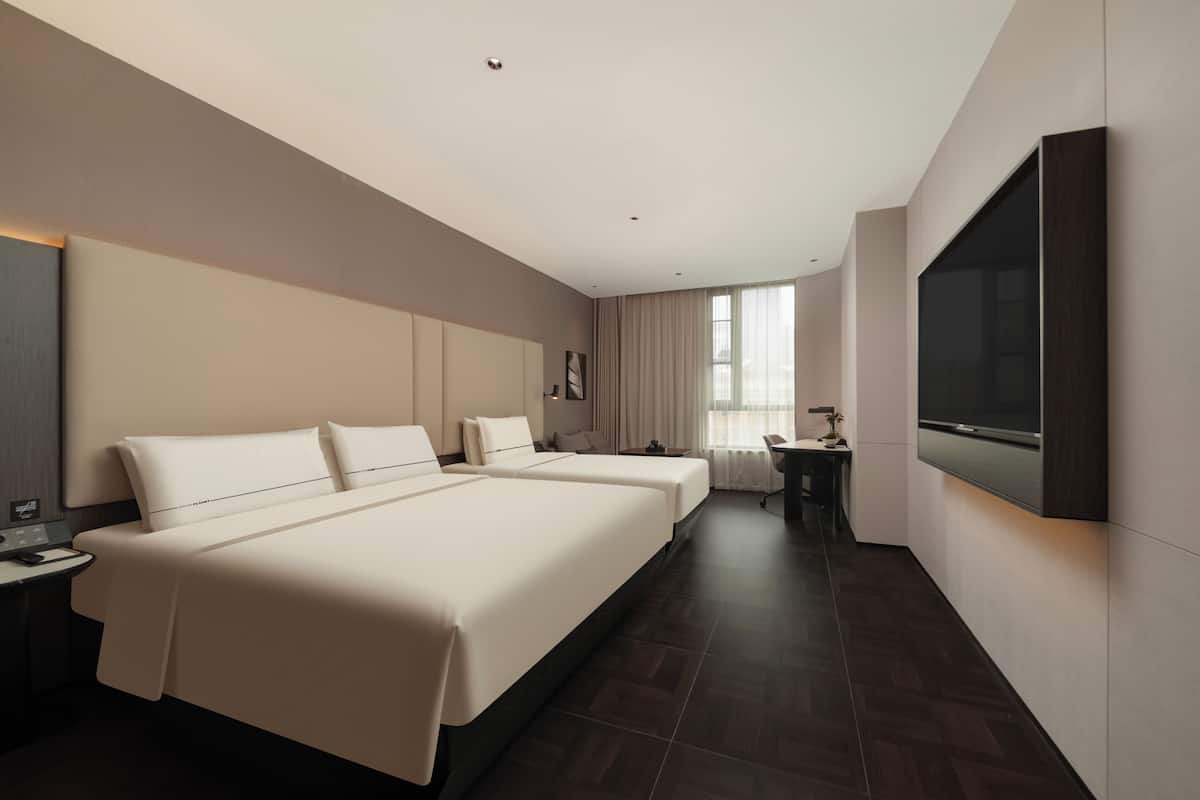 A modern hotel room with two beds, a desk, a chair, a flat-screen TV, and a large window.