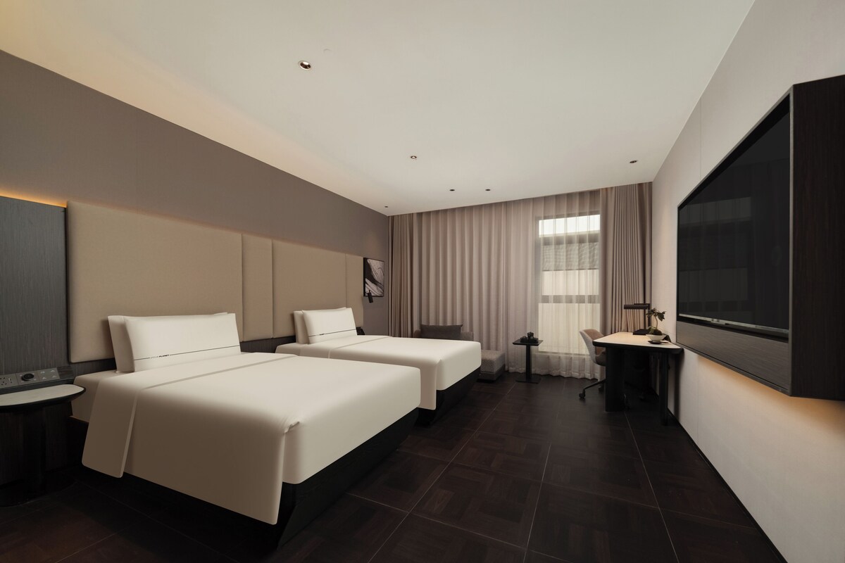 A hotel room with two beds, a desk, a chair, a TV, and a window with curtains.
