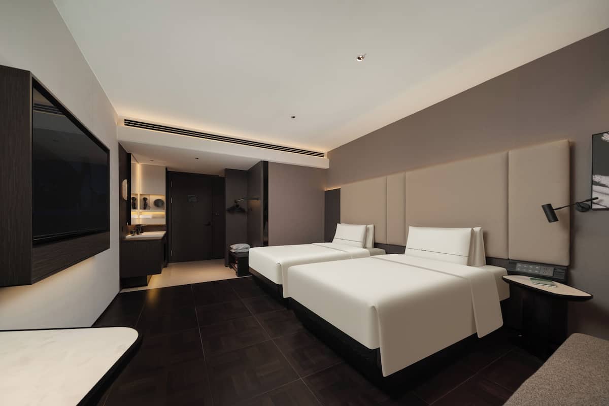 A modern hotel room with two beds, a desk, a vanity with a mirror, and a television.