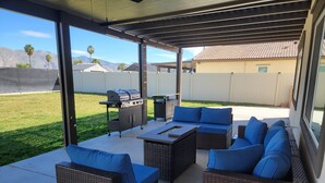 Un patio exterior con mobiliario de mimbre, una parrilla y vistas al jardín trasero.
