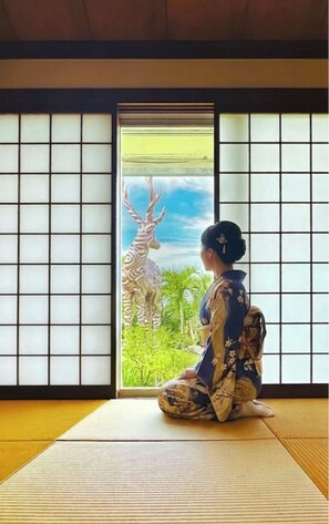 A room with tatami flooring, a sliding door with a view of greenery, and a person in traditional attire.