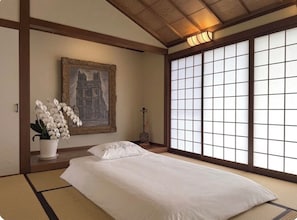 A traditional Japanese-style room with tatami flooring, a bed with white bedding, a framed picture of a building, and a shoji screen.