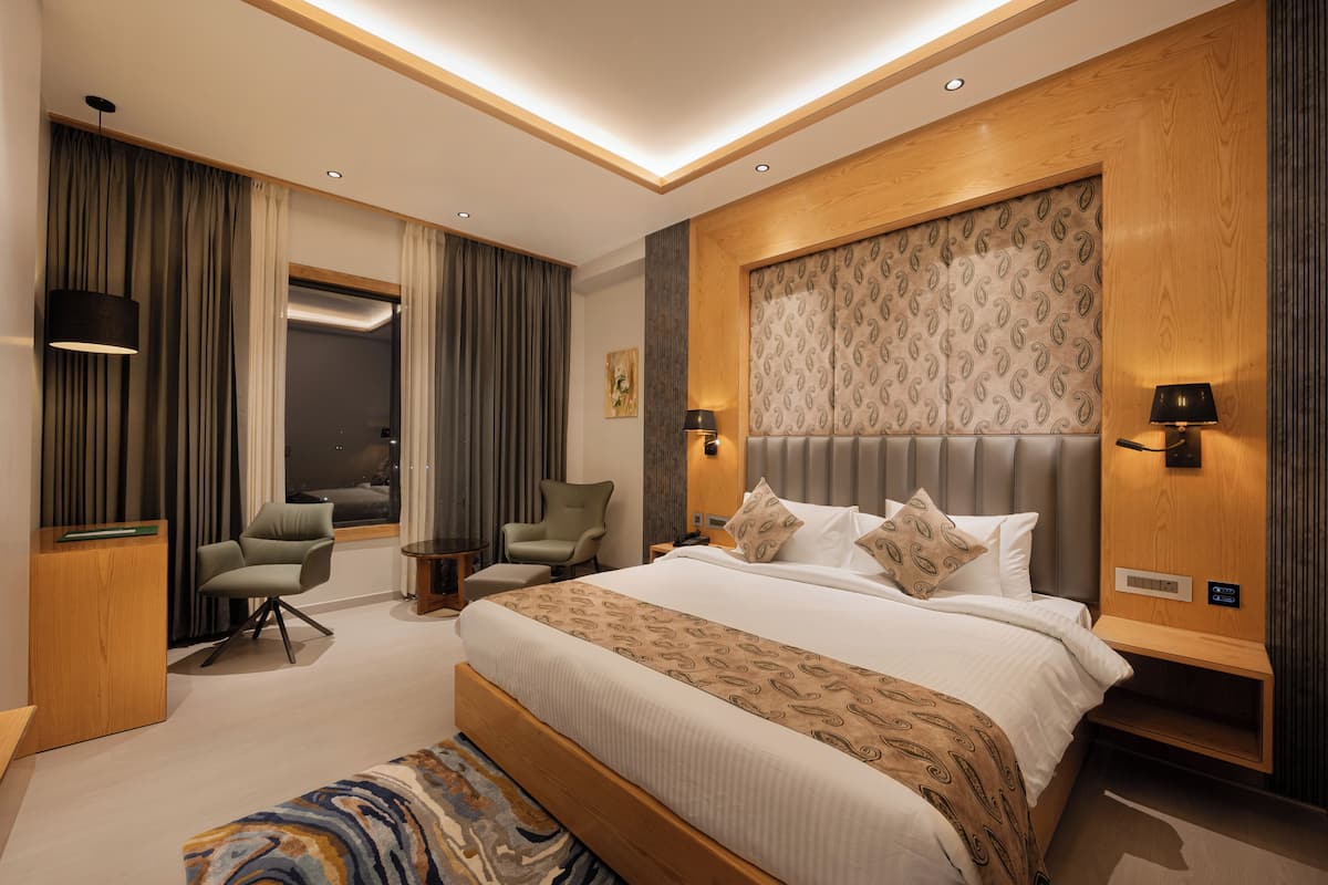 A modern hotel room with a large bed, a seating area with a chair and a small table, a desk, and a large window.