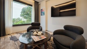 A modern room with a large window, a flat-screen TV mounted on the wall, two comfortable armchairs, a glass coffee table with a tray of tea and cups, and a patterned carpet. - Ananta Express (Katra)
