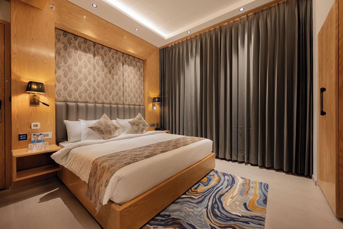 A modern hotel room with a large bed, wooden headboard, two bedside lamps, a nightstand, and a patterned curtain.