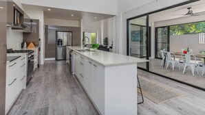 A modern kitchen with a central island, a dining area with white chairs, and a sliding glass door leading to an outdoor space. - Longboard Coastal Retreat (Kingscliff)