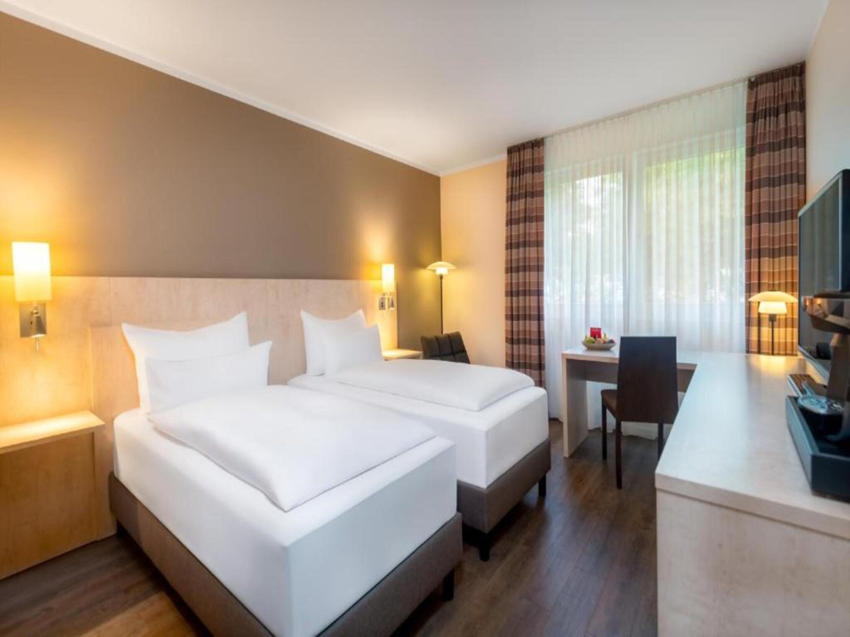 A hotel room with two beds, a desk, a chair, a TV, and a window with curtains.