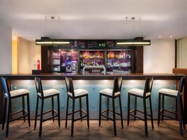 Una zona de bar con mostrador, taburetes y una variedad de botellas y vasos en estantes.