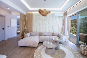 A modern living room with a sectional sofa, a round coffee table, and a view of a mountainous landscape through large glass doors. - Villa Oliva – Seaside Charm with Village at Your Doorstep (Lefkada)