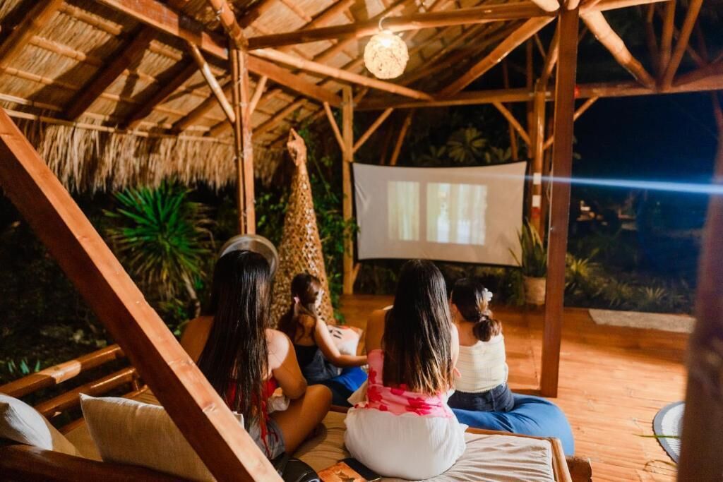 A wooden structure with a thatched roof, a projector screen, and people sitting on the floor.