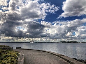 A waterfront promenade with a brick pathway, a body of water, and a cruise ship in the distance. - Heikendorf by Belvilla Single Room Land Side (Heikendorf)