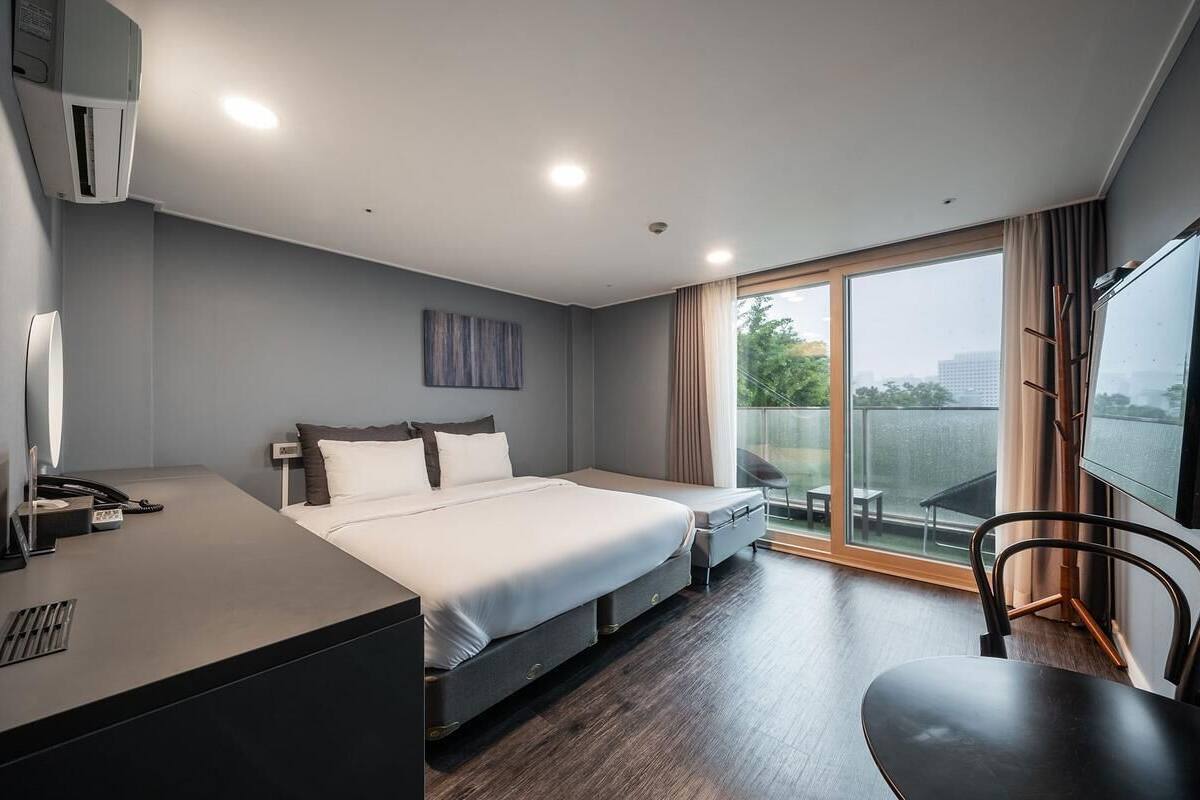 A hotel room with a bed, bedside table, desk, chair, TV, and a large window with a view.