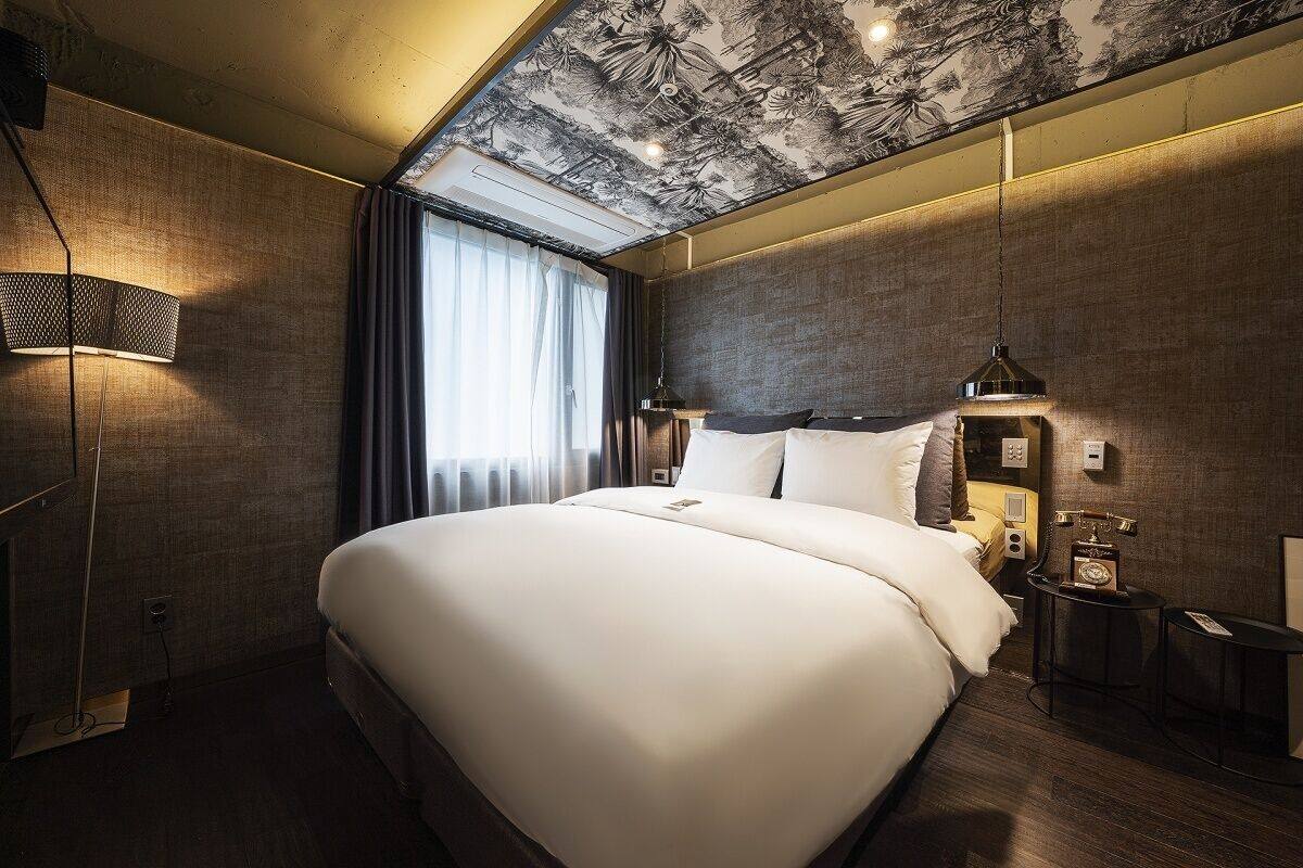 A modern hotel room with a large bed, a ceiling mural, a desk with a lamp, and a view of the outdoors.