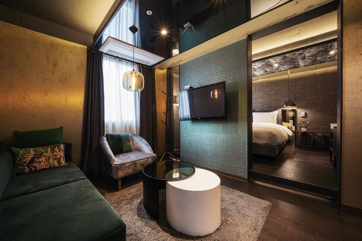 A modern hotel room with a sofa, a round coffee table, a TV, and a mirror reflecting the room.