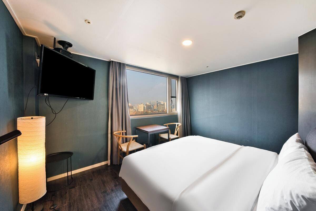 A hotel room with a bed, a TV mounted on the wall, a small table with chairs, and a window with a city view.