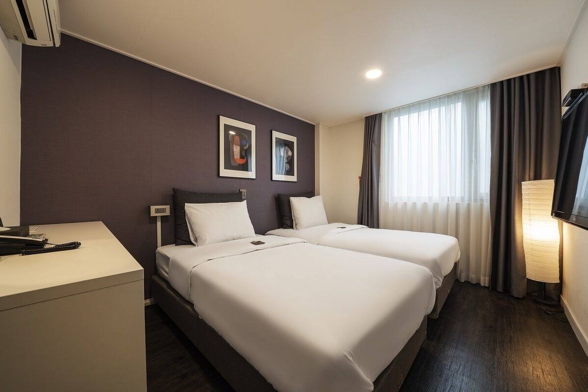 A hotel room with two beds, a desk, a TV, and a window with curtains.