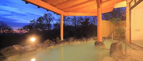 A hot spring area with a wooden pergola, rocks, and a view of trees at dusk.