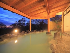 Un área de aguas termales con una pérgola de madera, rocas y vistas a árboles al anochecer.