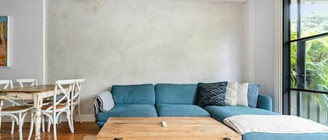A modern living room with a blue sofa, a wooden coffee table, and a dining area with white chairs.