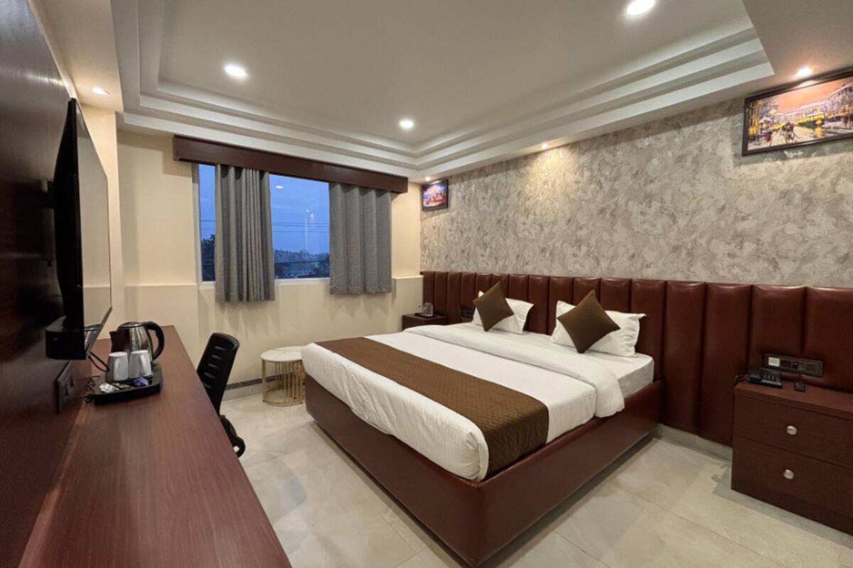 Quarto de hotel com uma cama grande, uma mesa de trabalho com cafeteira, televisão e janela com cortinas.