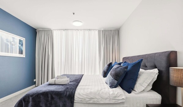 Hotellihuone, jossa on sänky, verhot, sängynpääty, yöpöytä ja lamppu.