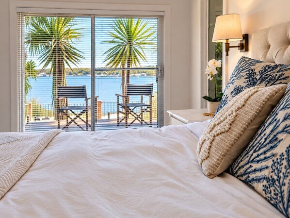 A bedroom with a large bed, a view of the sea through a sliding door, and a nightstand with a lamp.