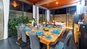 An outdoor dining area with a wooden table set for six, surrounded by chairs, with a kitchen and bar area visible in the background. - Melasti Beach Villa (Ungasan)