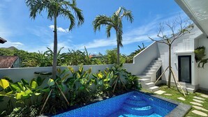 Piscina con borde alicatado, rodeada de exuberante vegetación y palmeras, con una escalera que lleva a la entrada de un edificio.