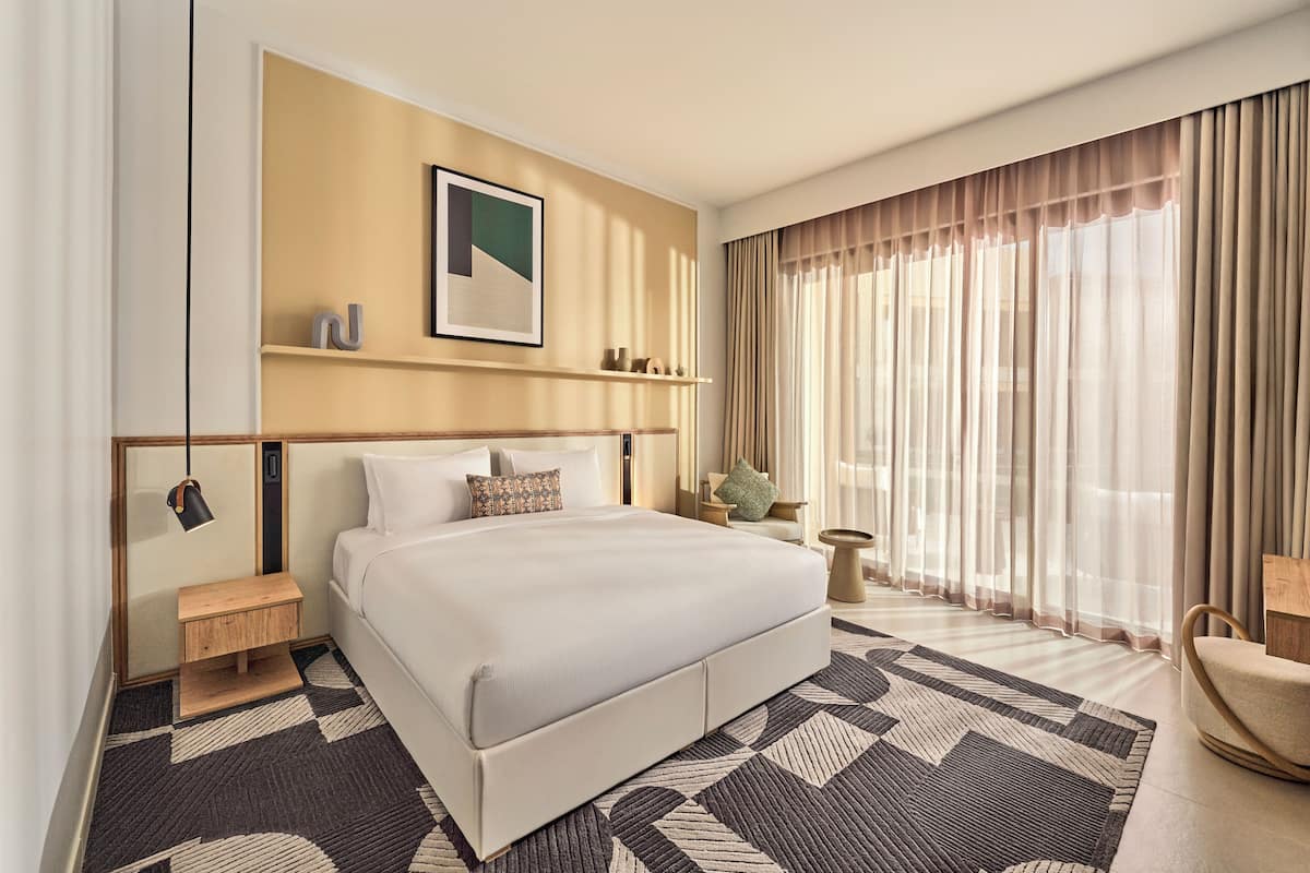 Habitación de hotel moderna con una cama grande, una mesita de noche, una silla y un ventanal con cortinas.
