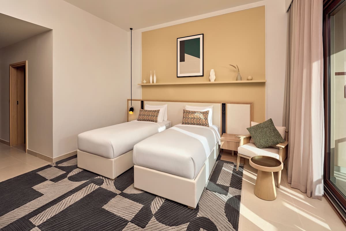 Habitación de hotel con dos camas, una mesita de noche, una lámpara, una silla, una lámpara de pie, un cuadro en la pared y una ventana con cortinas.