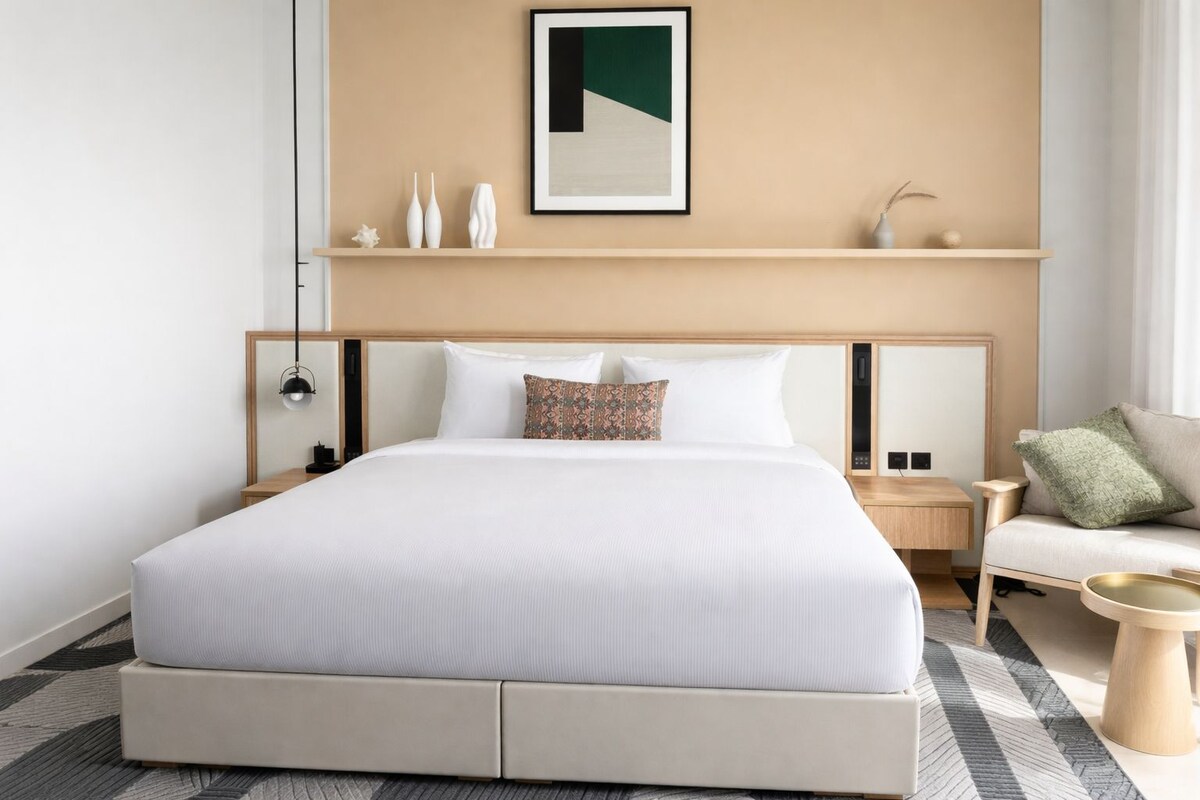 A modern hotel room with a large bed, a headboard, a bedside table, a nightstand, a sofa, and a wall-mounted shelf with decorative items.