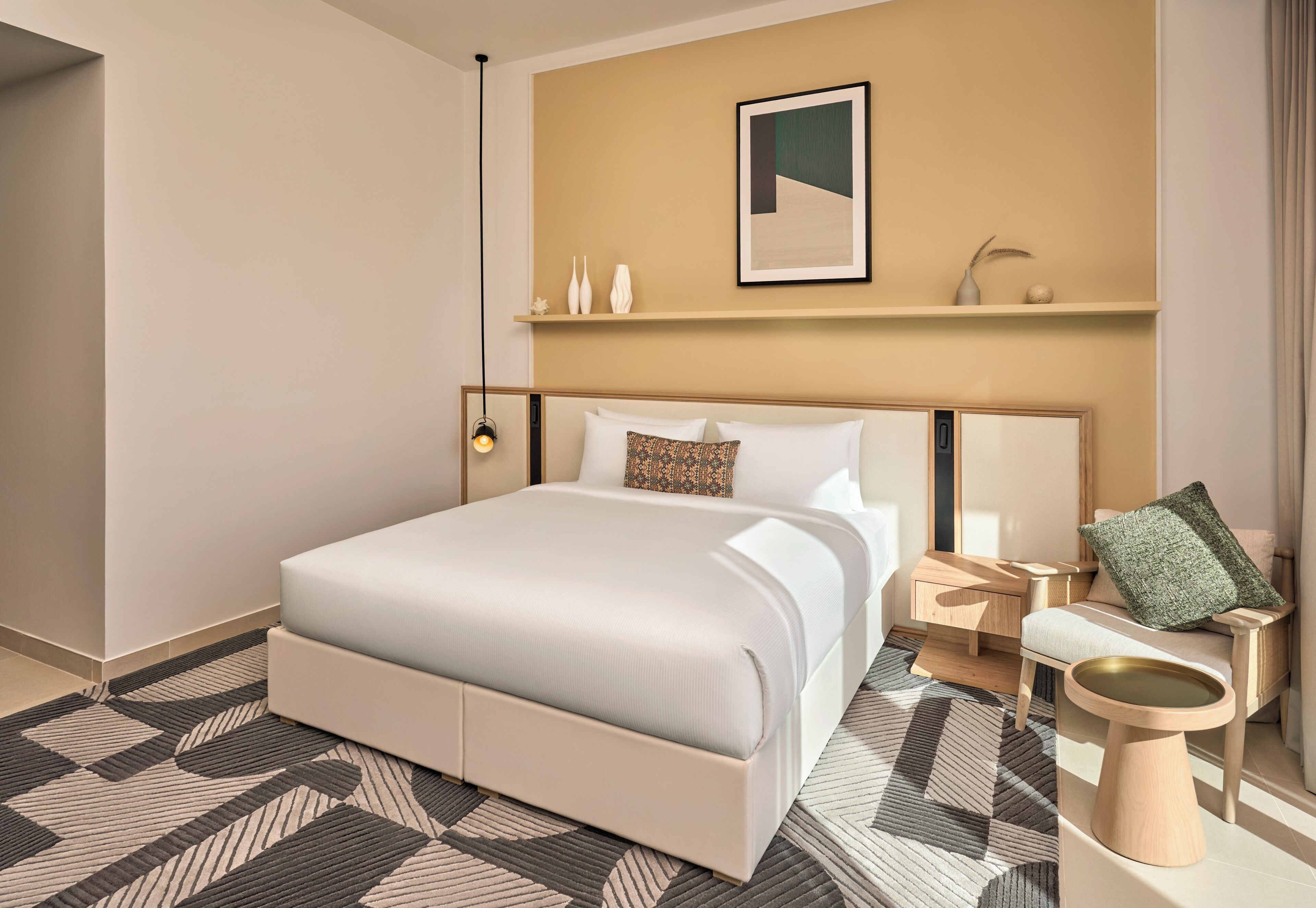 A modern hotel room with a large bed, bedside tables, a small side table, a wall-mounted shelf, and a framed picture.