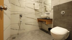 A bathroom with marble walls, a white toilet, a sink with a mirror, and a towel hanging on the wall. - Hotel Grace (New Delhi)