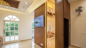 A hotel lobby with a large arched window, a flat-screen TV, a wooden partition, and a fan on the wall. - Hotel Grace (New Delhi)