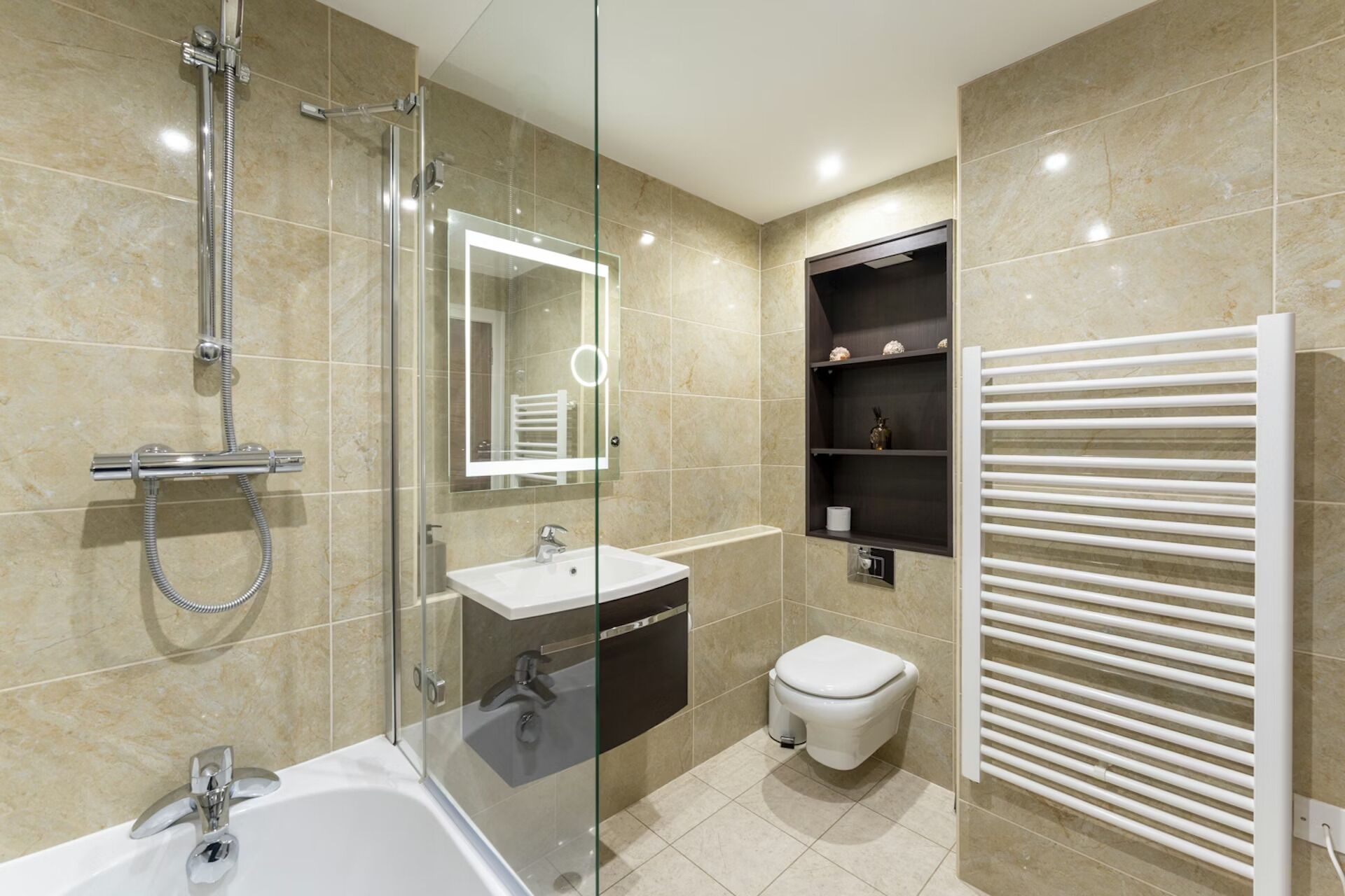 A modern bathroom with a glass-enclosed shower, a white bathtub, a sink, a toilet, and a built-in shelf with toiletries.