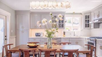 A kitchen with white cabinets, a dining table with chairs, a vase with flowers, and a hanging light fixture.
