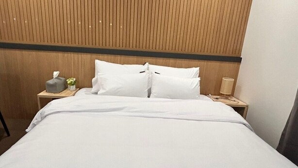 Una cama bien tendida con sábanas blancas, cabecera de madera, mesita de noche con una lámpara y una caja de pañuelos.