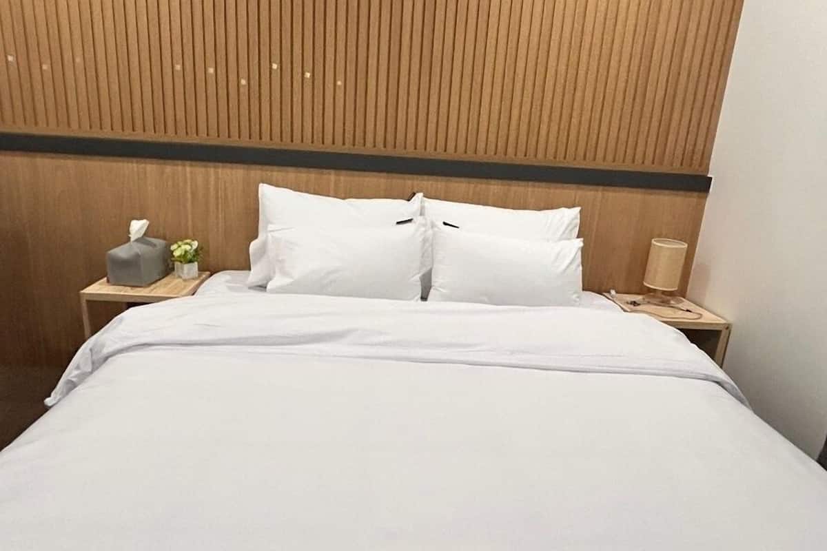 Una cama bien tendida con sábanas blancas, cabecera de madera, mesita de noche con una lámpara y una caja de pañuelos.