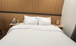 Een net opgemaakt bed met witte lakens, een houten hoofdbord, een nachtkastje met een lamp en een zakdoekendoos.