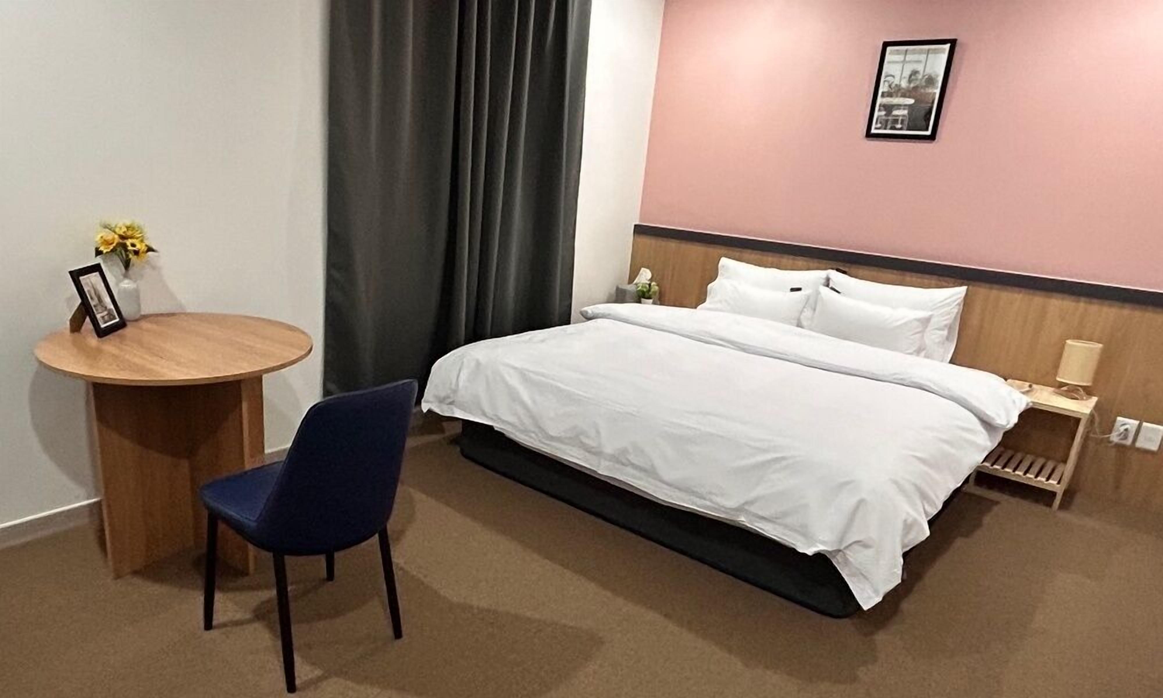 Una habitación de hotel con una cama, una mesa redonda de madera con una silla, una mesita de noche con una lámpara, y un cuadro en la pared.