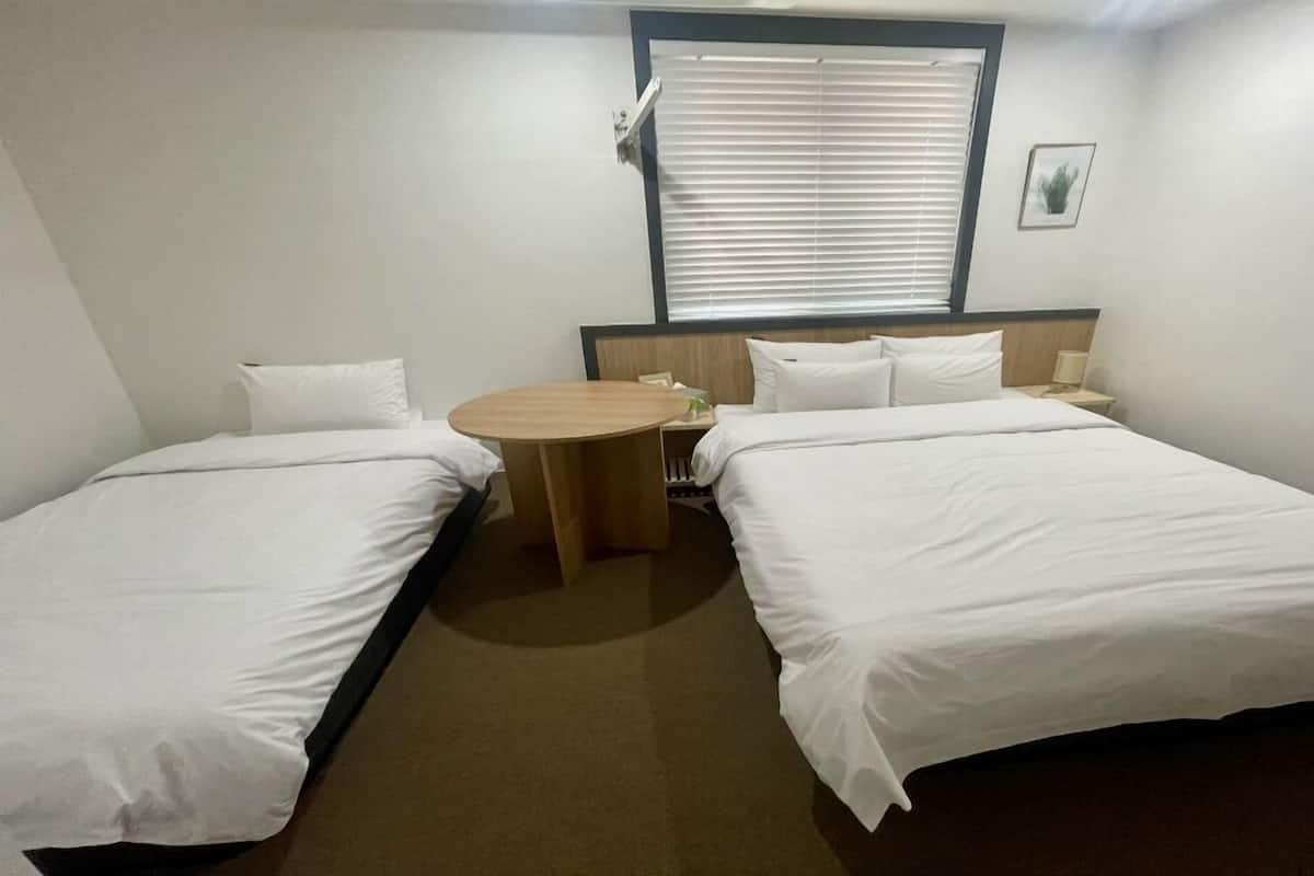 Habitación de hotel con dos camas individuales, una mesita redonda, una ventana con persianas y un cuadro en la pared.