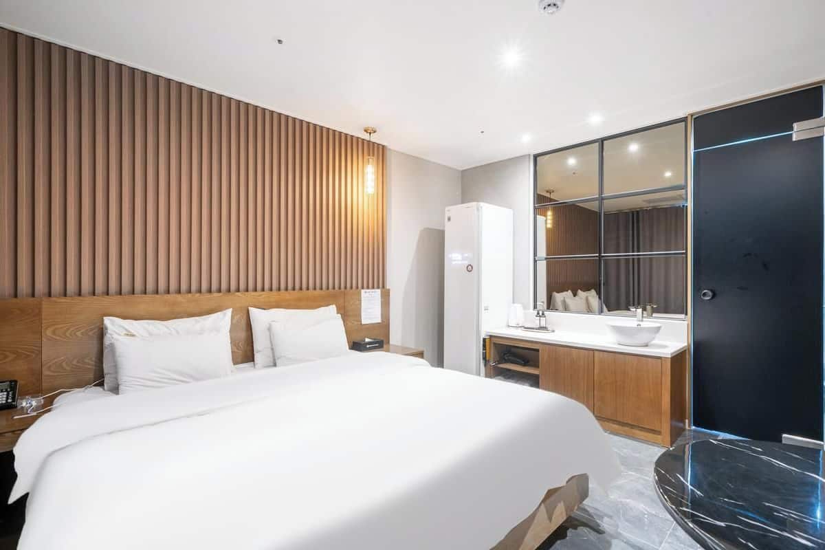 Habitación de hotel moderna con una cama grande, cabecera de madera, mesita de noche con teléfono, lámpara de noche blanca, baño con lavamanos, espejo y una puerta negra.